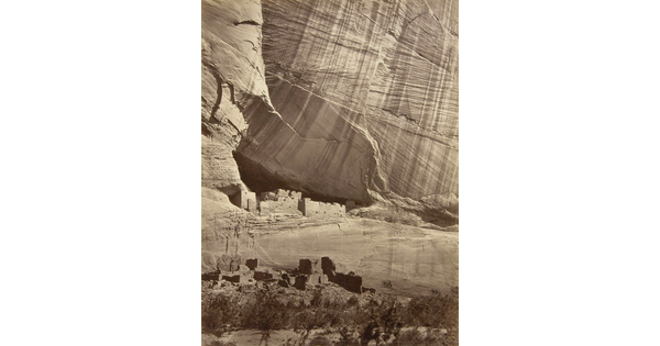 A sepia-toned photograph of cliff dwellings at the base of a large, sheer cliff.