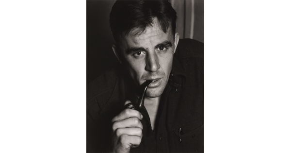 A black-and-white portrait photograph of a White man in a dark shirt leaning forward as he smokes a pipe.