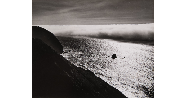 A black-and-white photograph taken from a hillside overlooking a calm ocean as thick clouds roll in or out.