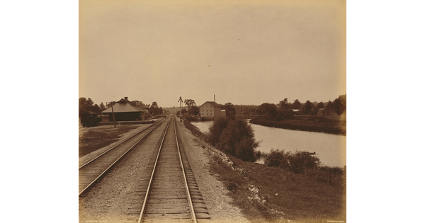 A sepia-toned photograph of railroad tracks leading to several buildings, and water to the right of the tracks.
