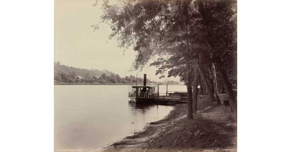 A black-and-white photograph of a boat docked on a river and trees on the shore.