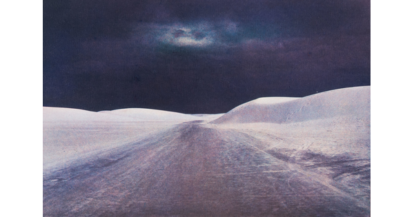 A color photograph of rolling white sand dunes on either side of a road under a dark and cloudy night sky.