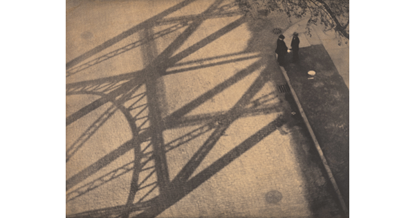 A black-and-white photograph taken from above of two people standing in the shadow of a bridge.