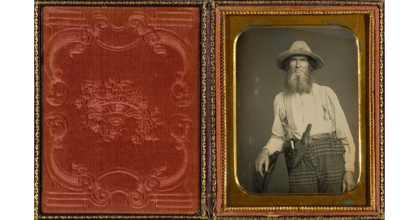 A black-and-white portrait photograph, in a gold frame and red velvet case, of a bearded White man wearing a hat, white shirt, and plaid pants with a knife tucked into the waistband.