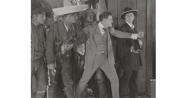 A black-and-white photograph of three medium-skinned men wearing sombreros and one White man in a suit restraining an angry White man in a three-piece suit.