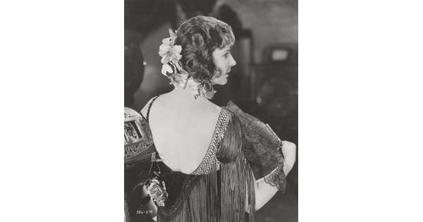 A black-and-white photograph, taken from the back, of a White woman wearing a low-back fringed frock, and fluttering a lacy fan.