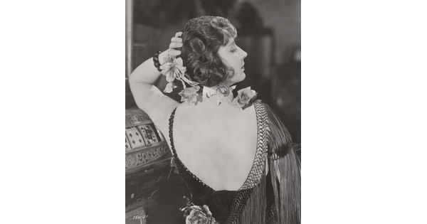 A black-and-white photograph, taken from the back, of a White woman, flowers in her hair, wearing a low-back fringed frock.