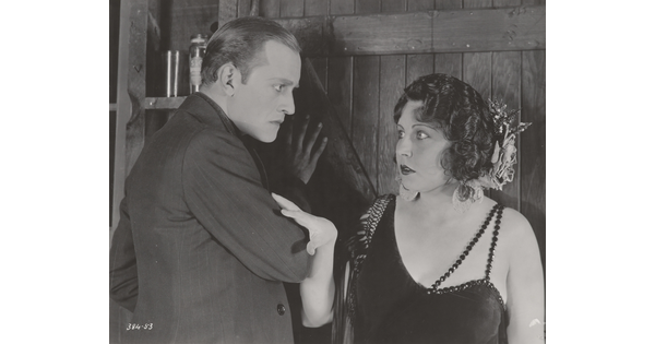 A black-and-white photograph of a White man looking intently at a White woman; she has her hand on his arm and looks behind him.