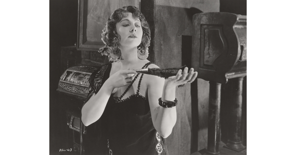 A black-and-white photograph of a White woman wearing a 1920s-style dress holding a feathered fan in her outstretched hands.