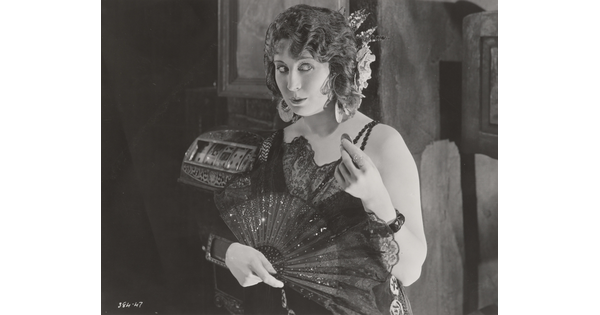 A black-and-white photograph of a White woman wearing a 1920s-style dress holding an open fan in one hand and a coin in the other.