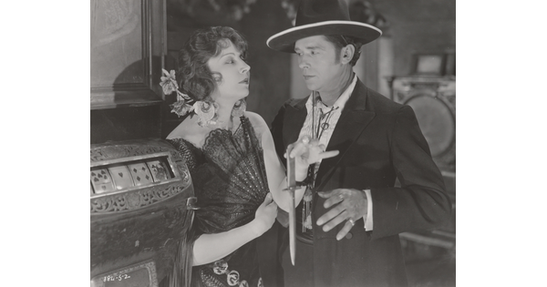 A black-and-white photograph of a White woman wearing a 1920s-style dress holding an open fan against her chest and a dagger as the White man next to her reaches for the dagger.