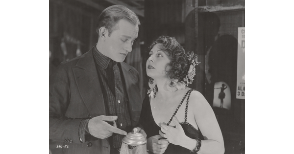 A black-and-white photograph of a White man and woman dressed in 1920s attire looking at each other as the man points to a coffee pot he is holding.