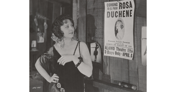 A black-and-white photograph of a White woman wearing a 1920s-style dress, one hand on her hip, looking at a poster nailed to a wall.