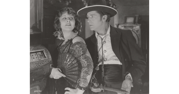 A black-and-white photograph of a White woman wearing a 1920s-style dress holding an open fan against her shoulder as the White man behind her holds a dagger.