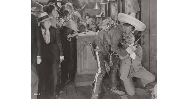 A black-and-white photograph of a group of people in a bar watching two men, one wearing a large sombrero and holding a revolver, wrestle over the gun.