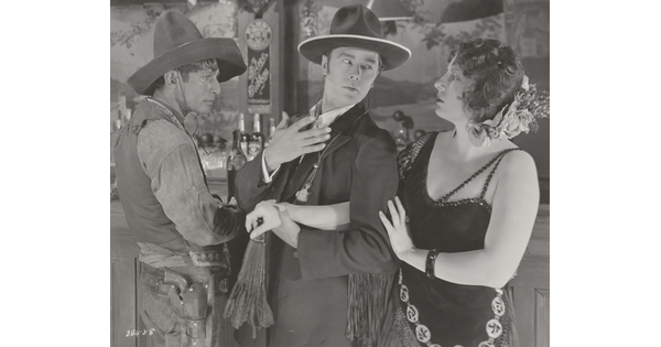 A black-and-white photograph of a medium-skinned man wearing a sombrero, a White man wearing a suit, and a White woman wearing a 1920s-style dress at a bar.