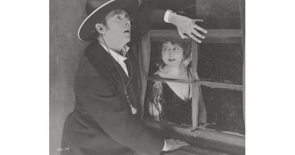 A black-and-white photograph of a White man standing next to a window, one hand under the window frame, while a White woman wearing a 1920s-style dress looks at him from the other side of the window.