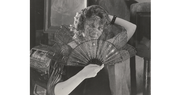 A black-and-white photograph of a White woman holding an open lace fan in front of her face.