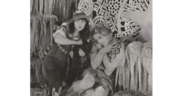 A black-and-white photograph of a White woman and man dressed in theatrical costumes in front of a set depicting snowflakes and icicles.