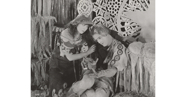 A black-and-white photograph of a White woman and man dressed in theatrical costumes in front of a set depicting snowflakes and icicles.