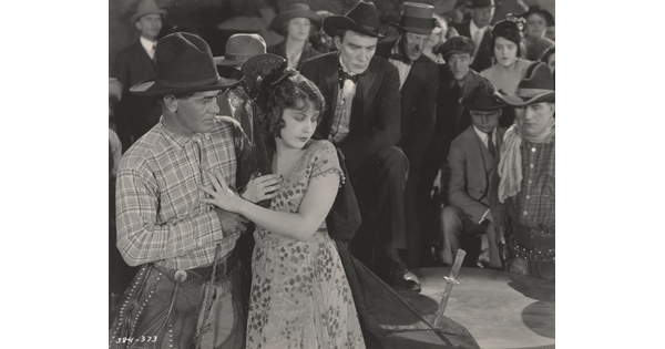 A black-and-white photograph of a White cowboy comforting a White woman whose veil is pinned to a table with a knife, as a crowd looks on behind her.