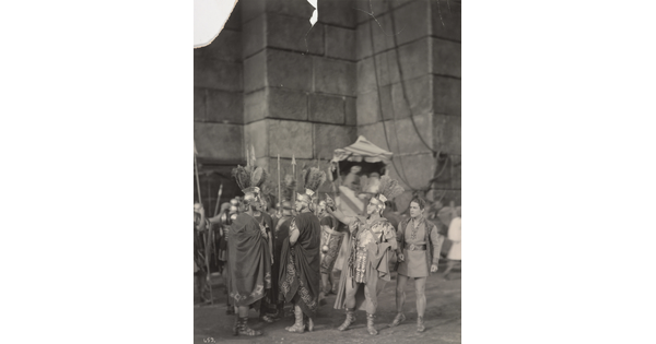 A black-and-white photograph of a group of people dressed as ancient Romans in front of a large stone building.