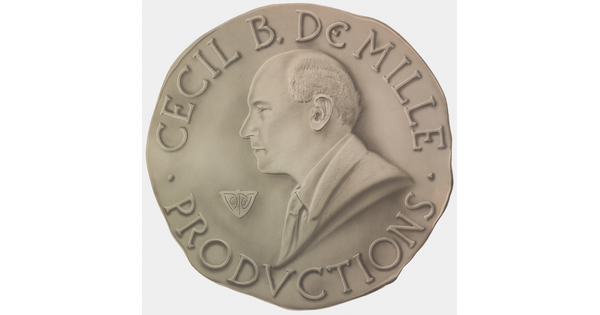 A sepia-toned photograph of a coin with a profile of a balding White man and the words "Cecil B. DeMille Productions" around the perimeter.