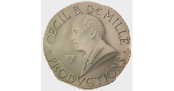 A sepia-toned photograph of a coin with a profile of a balding White man and the words "Cecil B. DeMille Productions" around the perimeter.