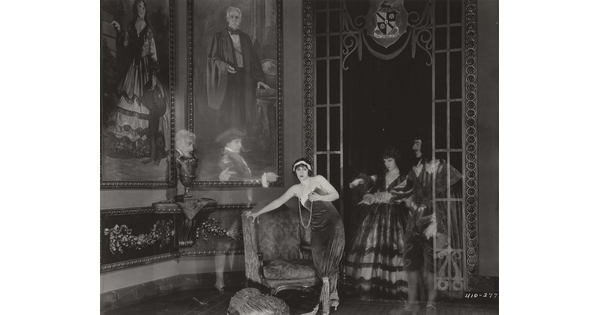 A black-and-white photograph of a White woman standing next to a loveseat in a lavish room as four transparent people in historical costumes point to her.