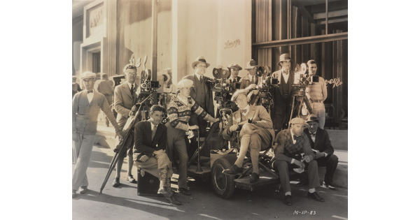 A black-and-white photograph of a group of men gathered outside of a building with film equipment.