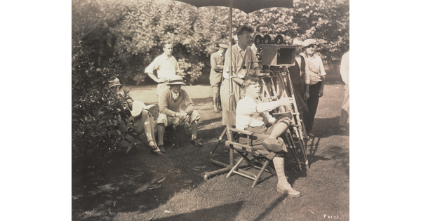 A black-and-white photograph of a group of White men, including a seated director gesturing in front of him, a camera man, and others in the background.