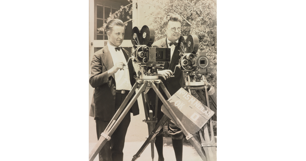 A black-and-white photograph of two White men standing next to each other behind two large video cameras.