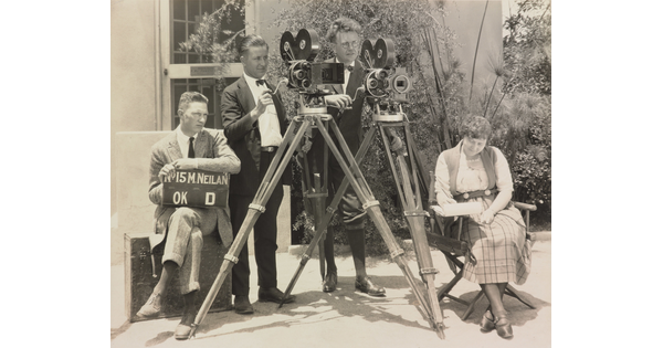 A black-and-white photograph of three White men, two standing behind video cameras, one seated with a clapperboard, and a White woman seated in a folding chair.