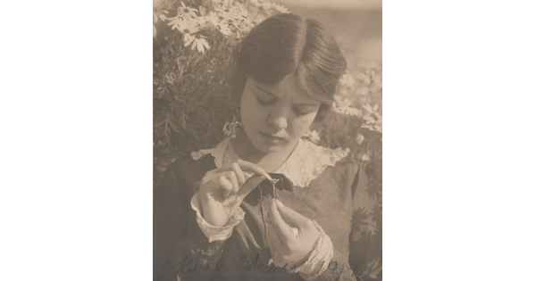 A black-and-white photograph of a young White woman picking the last petal off of a flower she holds in her hand.
