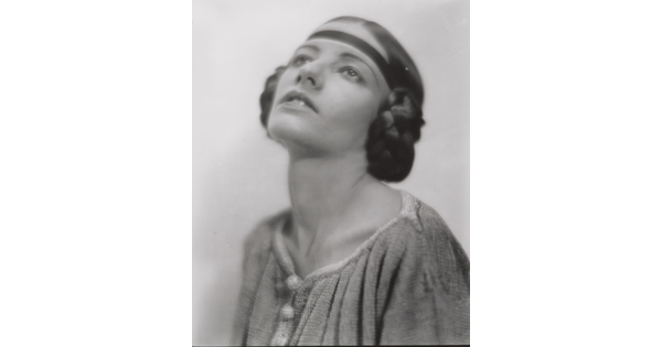 A black-and-white portrait of a White woman, head angled up, with her hair braided into buns on the sides of her head.