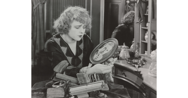 A black-and-white photograph of a White woman, seated at a vanity, gazing down at a photograph of a White man in an oval frame.