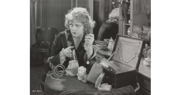 A black-and-white photograph of a White woman seated at a vanity, holding two handfuls of jewelry and looking off to the side.