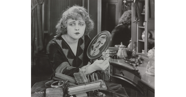 A black-and-white photograph of a White woman sitting at a vanity holding a brooch and a photograph of a White man as she gazes into the distance.