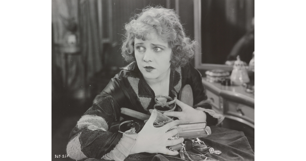 A black-and-white photograph of a White woman seated at a vanity clutching a variety of objects and jewelry to her chest.