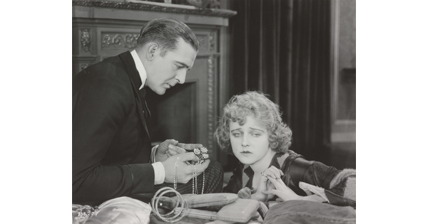 A black-and-white photograph of a White man presenting a White woman with jewelry as she sadly gazes into the distance.