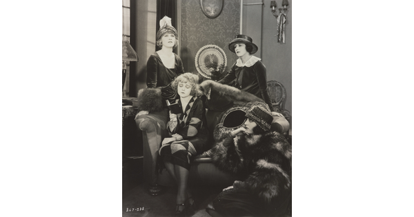 A black-and-white photograph of four elaborately-dressed White women, two standing, one reading on a sofa, the other seated on the floor.