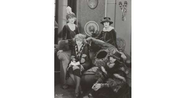 A black-and-white photograph of four elaborately-dressed White women, two standing, one on a sofa with a book in her lap, the other seated on the floor.