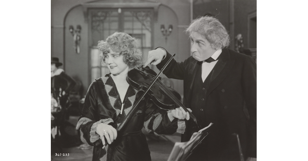 A black-and-white photograph of a White woman playing a violin and an older White man next to her with his finger violin's chinrest.