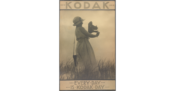 A sepia-toned photograph of a woman in a sunhat and long white dress standing in grass holding a large camera with text that reads, "Every day is Kodak day."