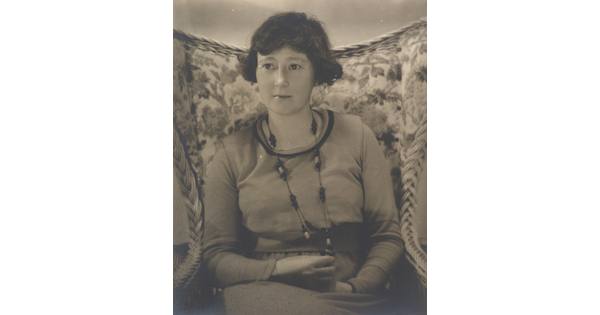 A black-and-white photograph a White woman with short dark hair wearing a long necklace and a long-sleeve top sitting in a floral, wingback chair.