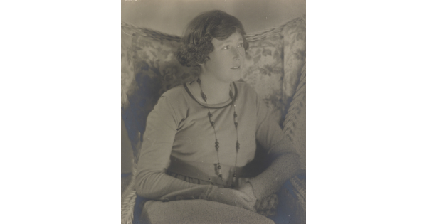 A black-and-white photograph a White woman, looking off to the side, with short dark hair wearing a long necklace and a long-sleeve top sitting in a floral, wingback chair.