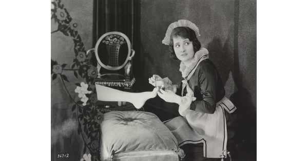 A black-and-white photograph of a White maid giving a pedicure to the foot of a woman who is hidden behind a screen.