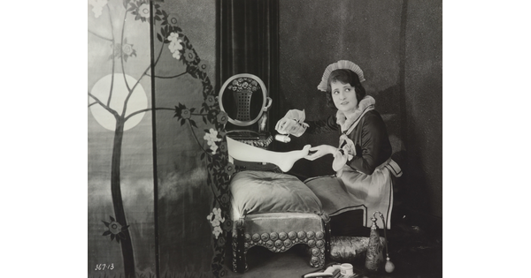 A black-and-white photograph of a White maid giving a pedicure to the foot of a woman who is hidden behind a screen.