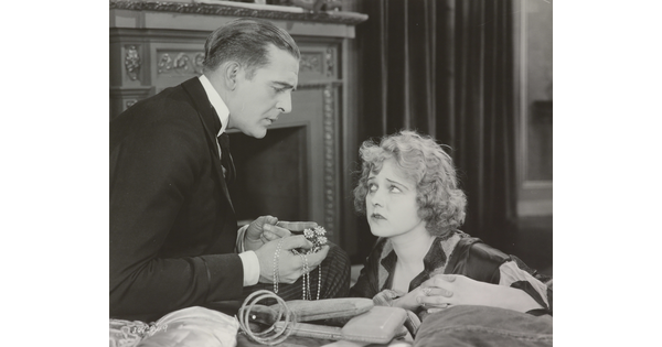 A black-and-white photograph of a White man presenting a White woman with jewelry as she gazes up at him looking worried.