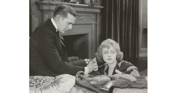 A black-and-white photograph of a White man presenting a White woman with jewelry as she sadly gazes into the distance.